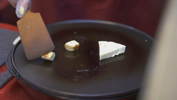 Hands close-up frying cheese in a frying pan. ART. The cook prepares cheese at the fair. Cook turns over tofu with a wooden spatula.