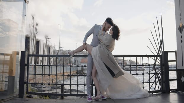 Beautiful couple of newlyweds posing by sea. Action. Attractive newlyweds pose on background of sea coast with city. Photo shoot of beautiful newlyweds in city by sea.