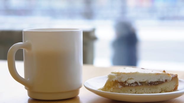Close-up of white mug and plate with cheesecake. Morning breakfast with coffee and classic cheesecake on wooden table. Sweet breakfast in cafe on background of window with busy street.