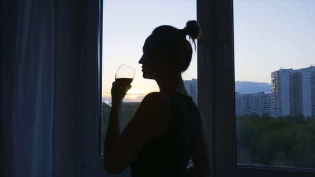 Woman drinks wine with pleasure on background of window. Action. Attractive woman drinks of red wine at home. Silhouette of woman drinking wine on background of evening window.
