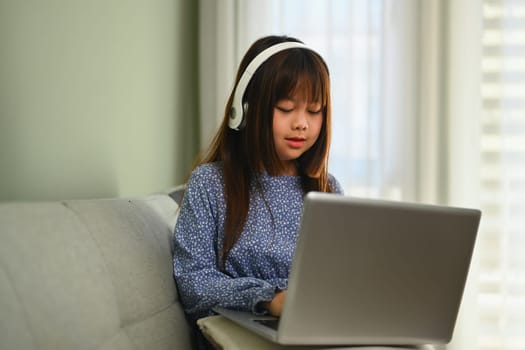 Concentrated Asian girl in headphone watching online lesson on laptop computer at home.