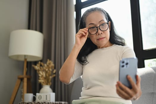 Beautiful middle age woman in glasses checking apps on mobile phone while sitting on couch.