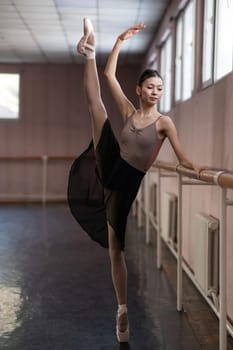 Graceful Asian ballerina in a beige bodysuit and black skirt is rehearsing in a dance class