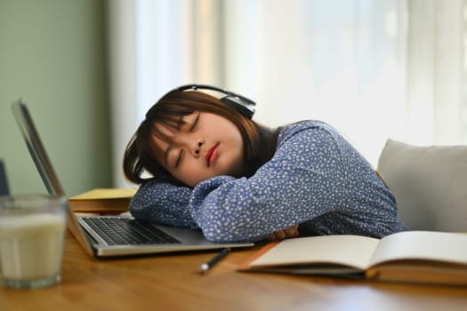 Tired Asian school girl sleeping on table while doing homework on laptop at home.