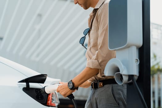 Young man put EV charger to recharge electric car's battery from charging station in city commercial parking lot. Rechargeable EV car for sustainable environmental friendly urban travel. Expedient