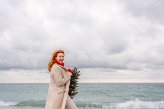 Redhead woman Christmas tree sea. Christmas portrait of a happy redhead woman walking along the beach and holding a Christmas tree in her hands. Dressed in a light coat, white suit and red mittens