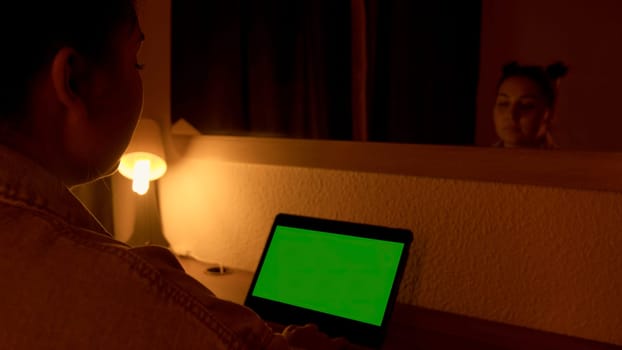 Woman sitting at the desk using laptop with green screen at night working over hours at home office. Media. Chroma key green screen for advertising
