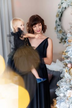 Mother daughter 2 years old Christmas tree. Both are dressed in black dresses, the mother holds the girl in her arms and both look at each other. The family celebrates Christmas