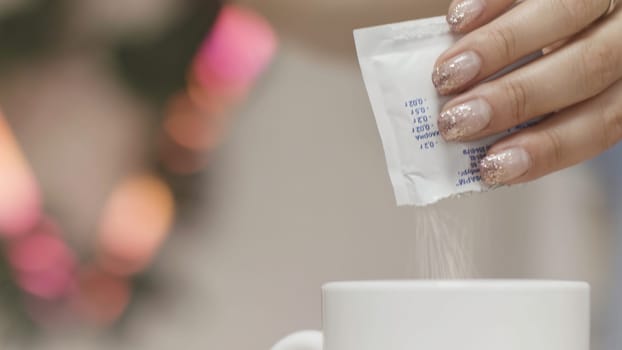 Close up of a woman hands throwing sugar into a white mug with hot beverage on blurred background. Female hands adding white sugar into tea or coffee.