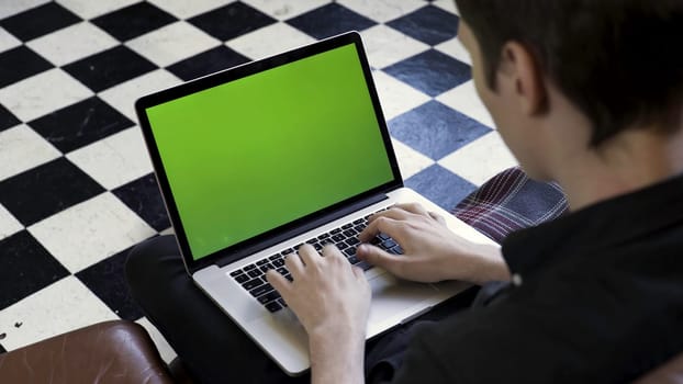 Over the shoulder view of a man using laptop with green screen in the room with chessboard floor. Male holding his device with chromakey screen and typing on it.