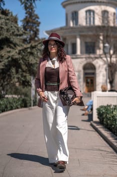 Woman park city. Stylish woman in a hat walks in a park in the city. Dressed in white corset trousers and a pink jacket with a bag in her hands