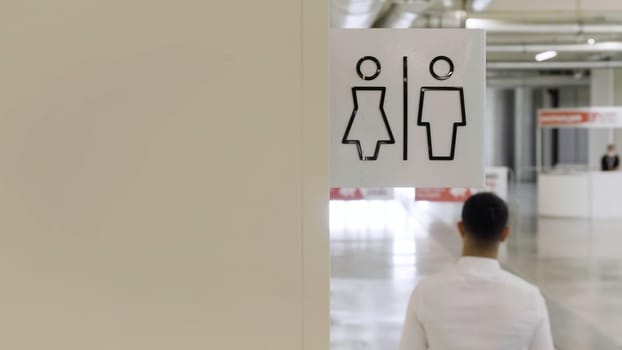 Men and women bathrooms sign at the shopping center. HDR. Rear view of a man in white shirt walking by the beige wall with male and female symbols of a public toilet