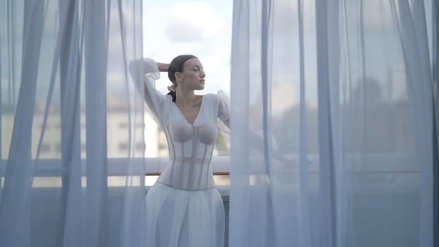 Beautiful woman posing in white dress with corset. Action. Elegant woman inwhite dress poses on terrace. Beautiful view through tulle of posing woman in white dress on terrace.
