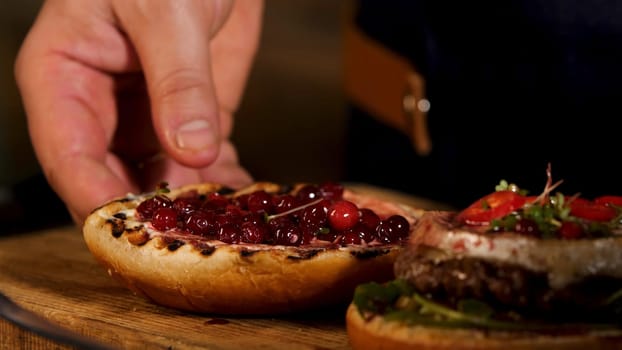 Close up of hamburger sandwich with cutlet of minced meat, cheese brie, camembert, berry cherry sauce. Male chef putting top burger bun on the other ingredients, foodporn and fast food concept.