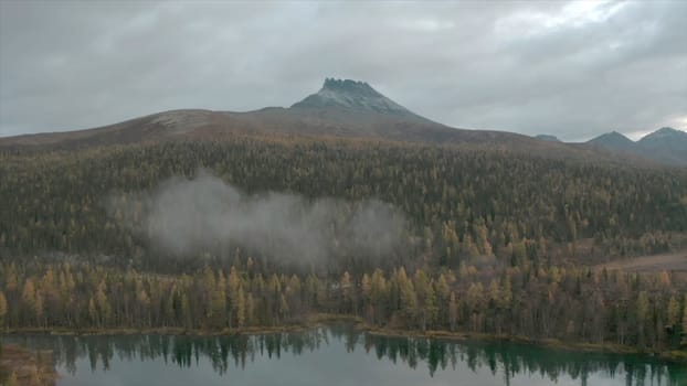 Top view of lake in forest part on background of mountain. Clip. Beautiful landscape with small lake in forest and mountain on horizon. Forest, lake and mountain on cloudy day.