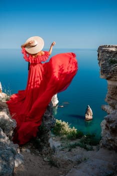 Red dress sea woman. Happy woman with flowing hair in a long flowing red dress stands on a rock near the sea. Travel concept, photo session at sea