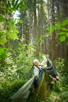 Adult mature woman 40-60 in a green long fairy dress in forest. Photo shoot in style of dryad and queen of nature. Fairy in beautiful green summer forest. Concept of caring for nature