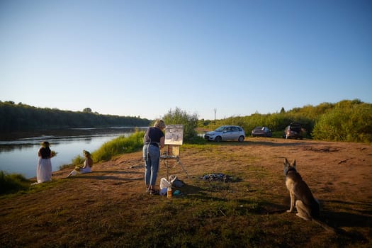 Adult female artist painting picture near water of river or lake in nature and girls in white sundress and flower wreath. Artist and models posing in holiday of Ivan Kupala in nature at sunset