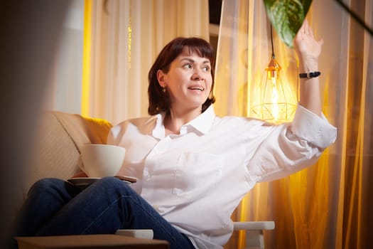 Nice girl with a mug of tea or coffee in cozy room. A middle-aged adult woman relaxing in the evening in the living room or bedroom in the evening