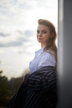 Portrait of Beautiful girl with red hair in white shirt in open wooden pavillion in village or small town. Young slender woman and sky with clouds on background on autumn, spring or summer evening