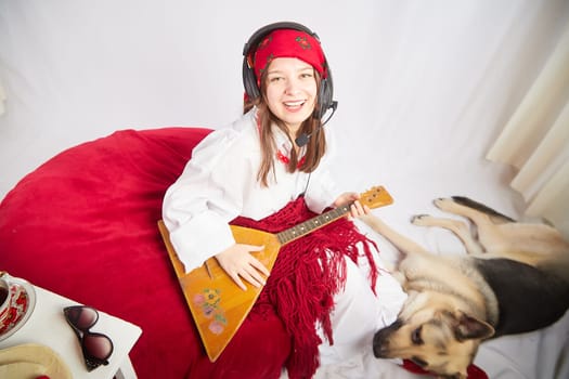 A girl in red scarf, white shirt, with shawl and balalaika in black sunglasses with a shepherd dog. Cheerful radio presenter with headphones and a microphone sings and has fun with an animal friend