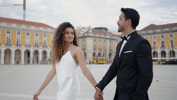 Young and beautiful couple walking along historic buildings and talking. Action. Man in suit and woman in white dress holding hands in the city square