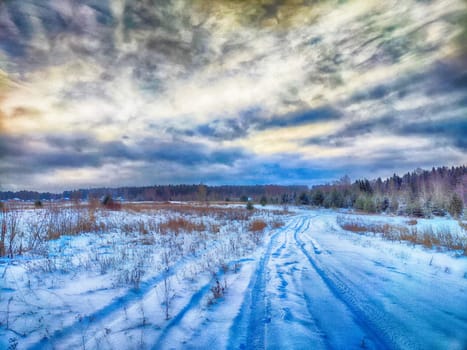 Dramatic sky with grey and blue clouds and field with snow and a rutted road in winter or autumn. Natural landscape. Dramatic sky sunset. Beautiful winter landscape