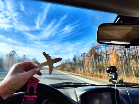 Small wooden toy airplane in the female driver's hand in car on the background of the road and highway. The concept of choosing the type of transport to travel. Choosing between an airplane and car