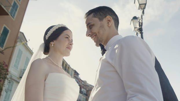 Bride and groom walking in a city street in summer day. Action. Low angle view of a woman with decoration on her hair and a veil standing near her groom in white shirt against the sun