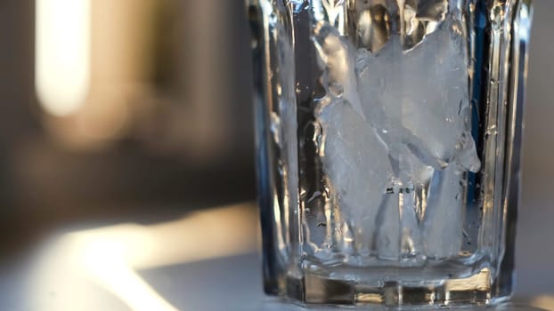 Throwing ice pieces into a transparent glass, preparing cocktail. Close up of crystal glass standing on the table filled with cold ice on blurred background.