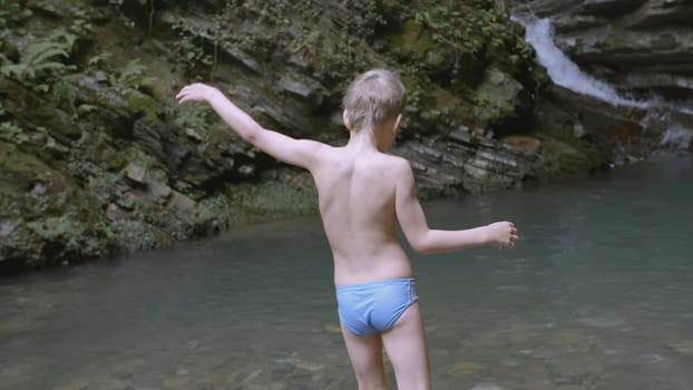 The boy enters the water near the waterfall to swim. CREATIVE. A white child in blue shorts enters the water. In the mountains there is a guy waist-deep in water.