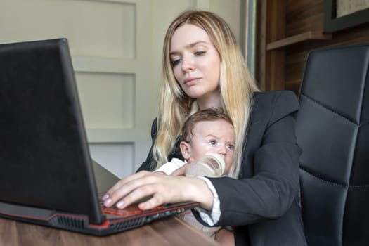 Concept of combining business and caring for newborn baby: young businesswoman holds baby in her arms, in office, combining this with working on laptop