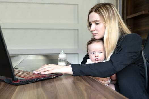 Young woman holds baby in her arms, in office, combining this with working on laptop., concept of combining business and caring for newborn baby