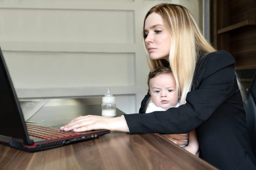 Concept of combining business and caring for newborn baby: young businesswoman holds baby in her arms, in office, combining this with working on laptop.