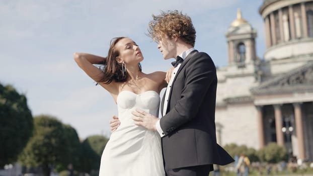 Beautiful newlyweds on background of old architecture. Action. Elegant couple of newlyweds posing on background of cathedral on sunny day. Newlyweds on windy sunny day in summer.