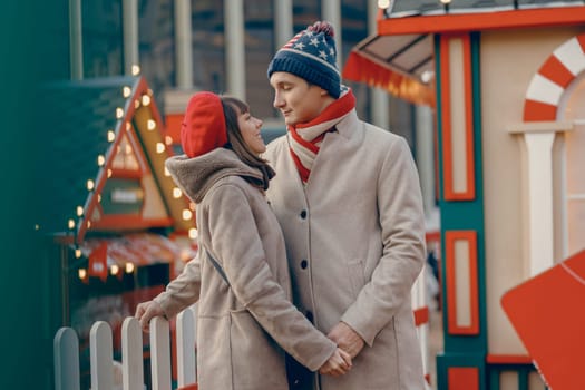 Surrounded by holiday lights, a couple in cozy winter hats finds romance in a loving gaze at the Christmas fair