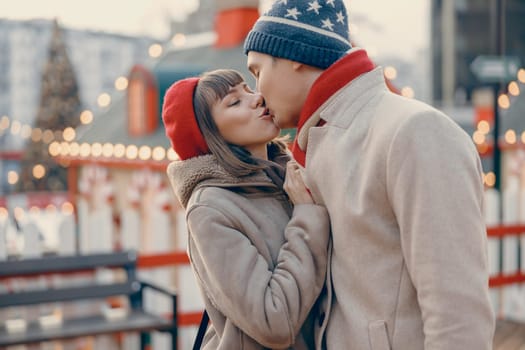 A tender moment as a young couple in winter attire shares a loving kiss amidst the twinkling lights of a Christmas market.