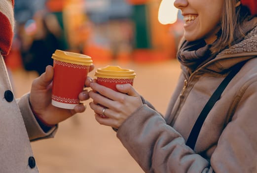Wrapped in winter layers, a couple laughs and holds warm drinks in holiday-themed cups, enjoying the festive cheer of a Christmas market