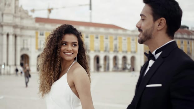 Young and beautiful couple walking along historic buildings and talking. Action. Man in suit and woman in white dress holding hands in the city square