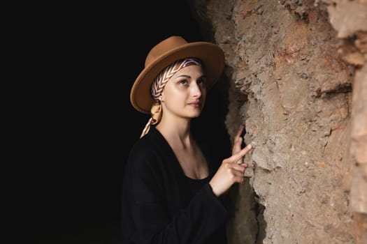 Woman in hat stands and looking at the wall of the cave in the settlement of ancient people. archaeological tourism concept.