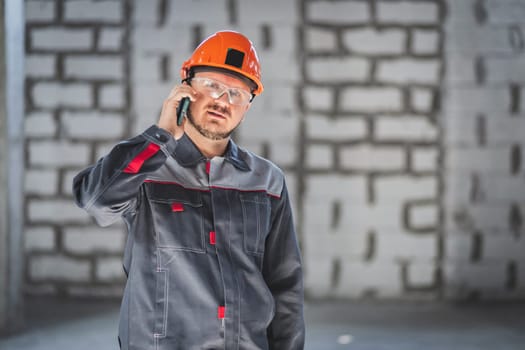 Bearded construction caucasian worker in boilersuit, helmet and protective glusses talking on a smartphone at a construction site.