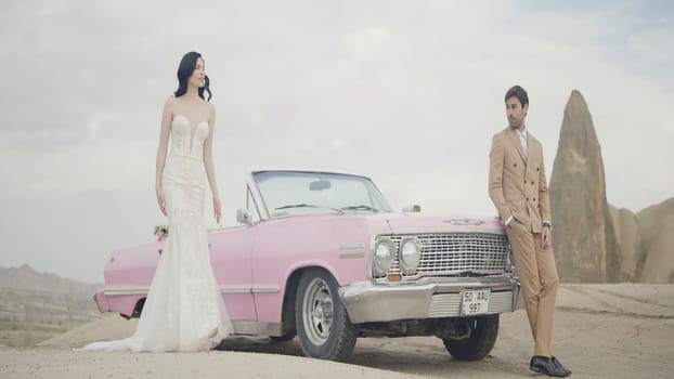 Beautiful outdoor shot next to a pink car. Action. An elegant couple with a brunette girl in a white dress and a man in a suit are walking in front of a pink car. High quality 4k footage