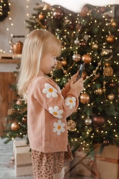 A little blonde girl photographs balloons on a Christmas tree in a festive interior decorated in a New Year's style. The concept of a merry Christmas