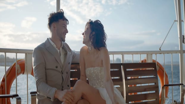 Wedding couple sitting on a bench outdoors in front of the sea. Action. Young and beautiful man and woman on a windy day