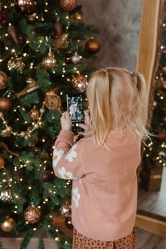 A little blonde girl photographs balloons on a Christmas tree in a festive interior decorated in a New Year's style. The concept of a merry Christmas