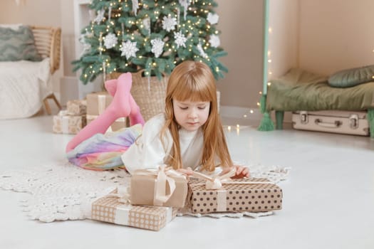 A little red-haired girl lies next to Christmas gifts in a craft package. Happy Christmas concept