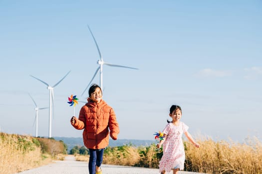 Two children girl with pinwheel happily near windmills. Family moments at turbines symbolize clean energy. Children joy windmill circle in sky represents sustainable cheerful community.