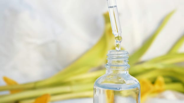 Vial with essential oil and yellow soft petals isolated on a white textile background. Girl hand touching cosmetic package surrounded by tulip leaves.