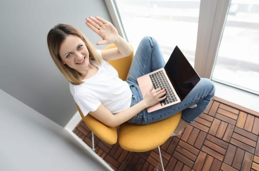 Young woman sitting on armchair with laptop and waving hand. Stay home concept