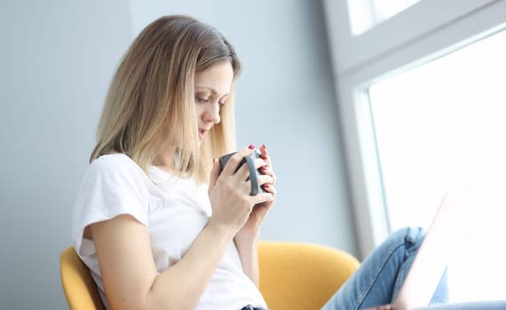 Woman with cup of coffee sitting on armchair with laptop. Self-isolation during covid19 pandemic concept